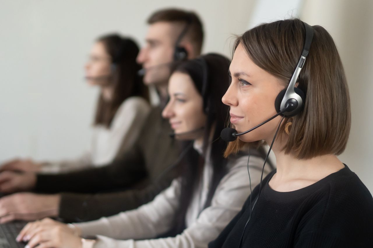 Side view of young coworkers in casual clothes sitting in light workplace with headphones with microphone while surfing on laptop